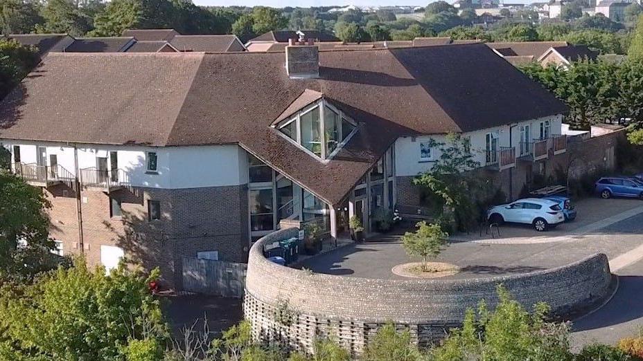 An elevated image of the Sussex Beacon facility on Bevendean Road in
Brighton. The centre has a circular area for cars to drop off and pick up visitors to the facility, which has a diamond window above the entrance doors.