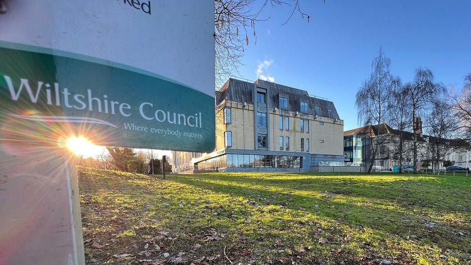 The bottom of a Wiltshire Council sign showing the logo with part of its HQ in the background on a sunny day with blue sky.