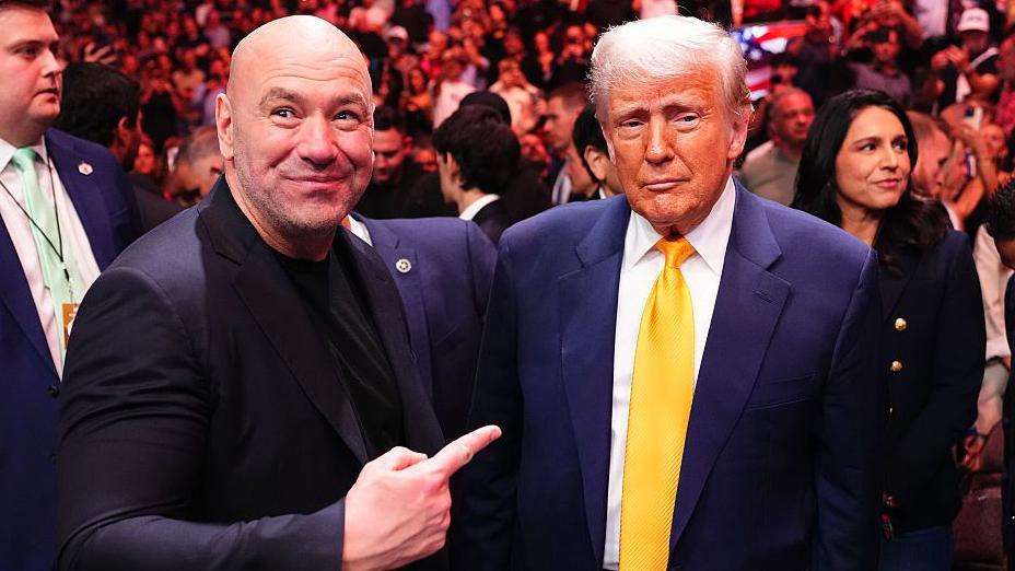 Dana White points and smiles at Donald Trump while standing shoulder to shoulder with him at a UFC event. Both are wearing suits and there is a large crowd in the background.
