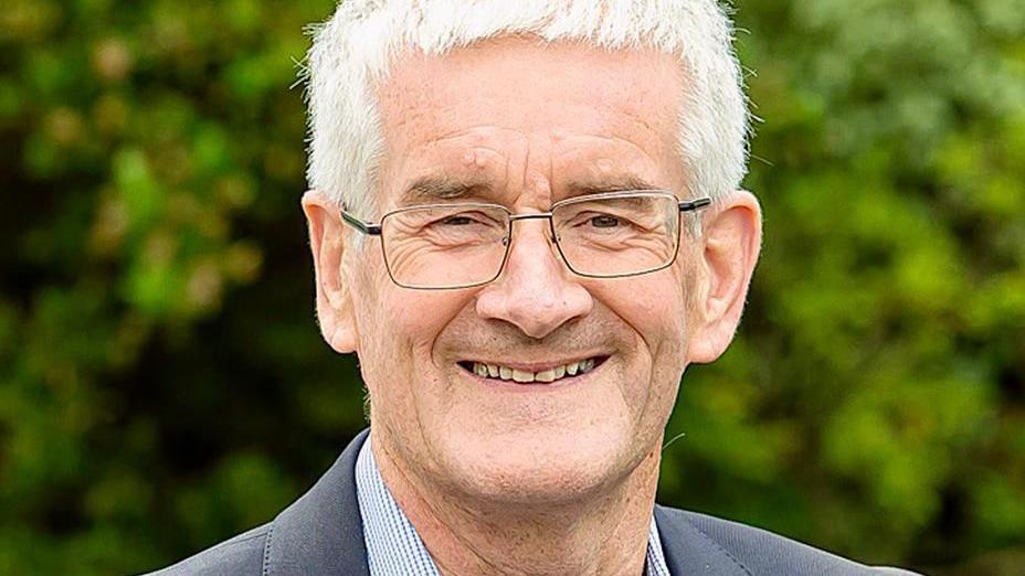 A head-and-shoulders photo of a man with white hair and glasses, smiling at the camera. There's some greenery in soft focus behind him.
