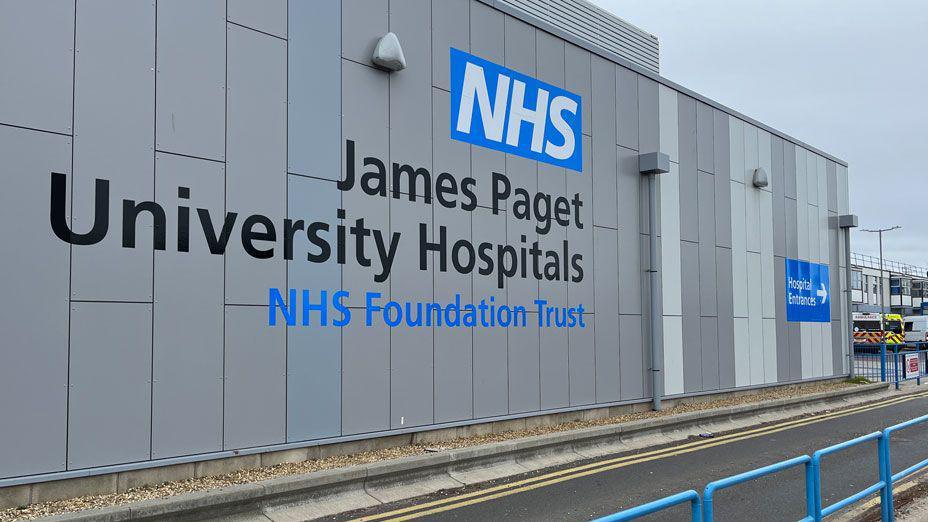 A grey wall of a hospital which says NHS James Paget University Hospitals and in front of it a road. Beyond it to the far right can be seen two ambulances