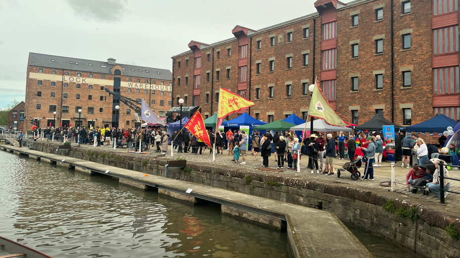Many people are standing on the edge of Gloucester Docks, standing in between large flags and red brick buildings.