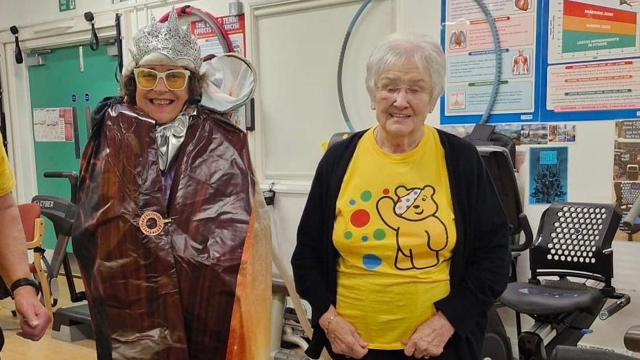 Three people standing in a gym in front of weight lifting machines. The woman in the middle is wearing a fancy dress costume that incorporates The Lion, The Witch and The Wardrobe from the C.S Lewis novel. The two people beside her are wearing yellow Children in Need tops with Pudsey Bear on.