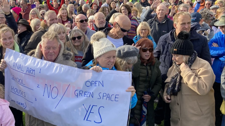 A group of people standing together, with a couple at the front holding a white banner which reads: Canal Arm 1 School 2 Houses 3 NO / Open Green Space Yes 