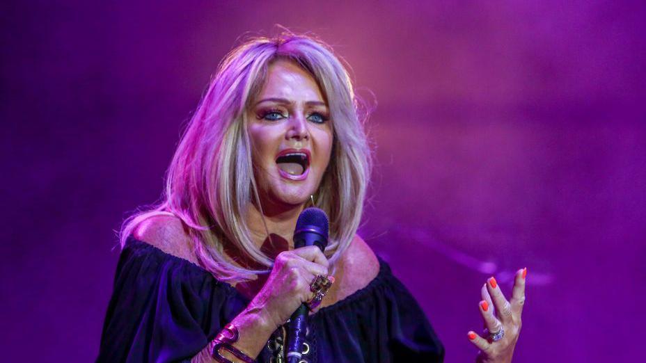 Bonnie Tyler on stage in purple pink lighting wearing black top and holding microphone. She has long blonde hair worn down.
