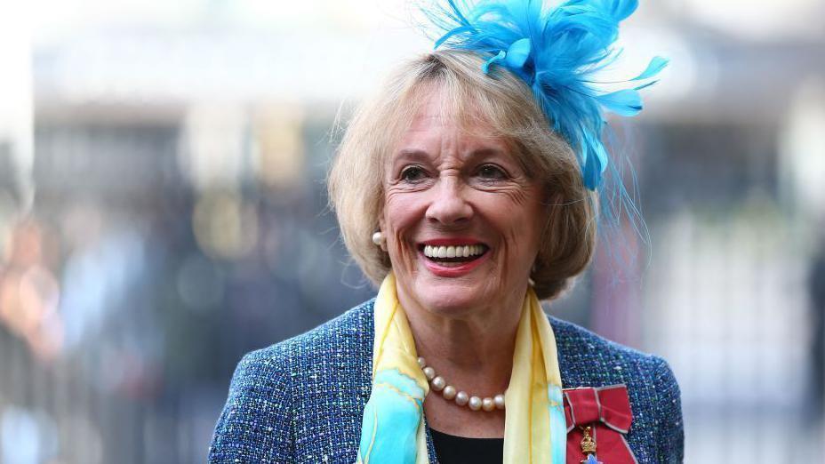 Dame Esther Rantzen looks slightly off camera, smiling. She has short blonde hair with a fringe framing her face and wears large pearl stud earrings and a single row of pearls as a necklace to match. She is wearing a blue feather fascinator on her head, with a blue tweed jacket and a yellow and pale blue silk scarf around her neck and black top underneath. She has a red bow with a medal pinned to the left panel of her jacket - a symbol of her damehood.