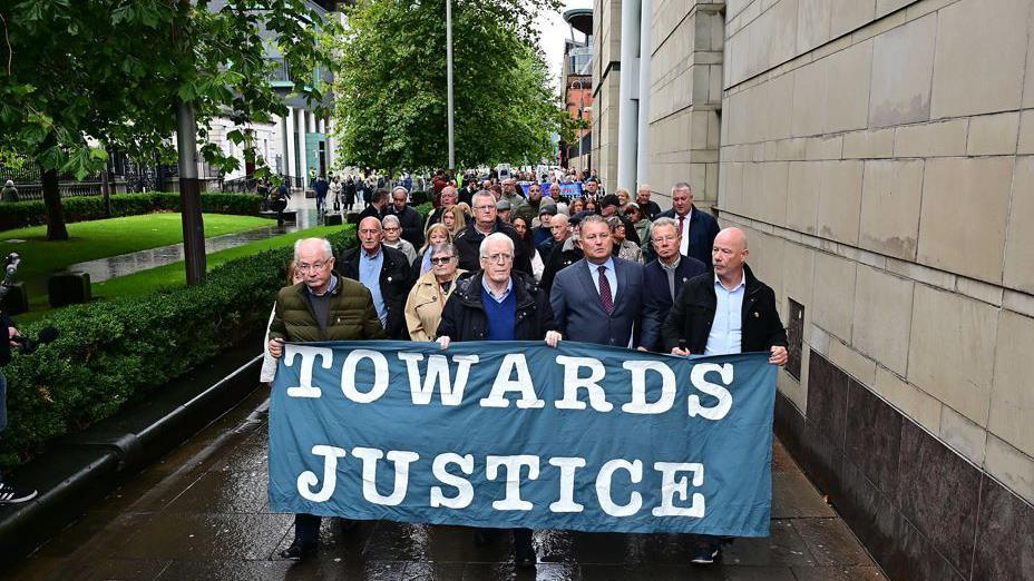 A crowd of people walk down a street holding placards and a banner that reads 'Towards Justice'. There are trees lining the street. The sky is grey.