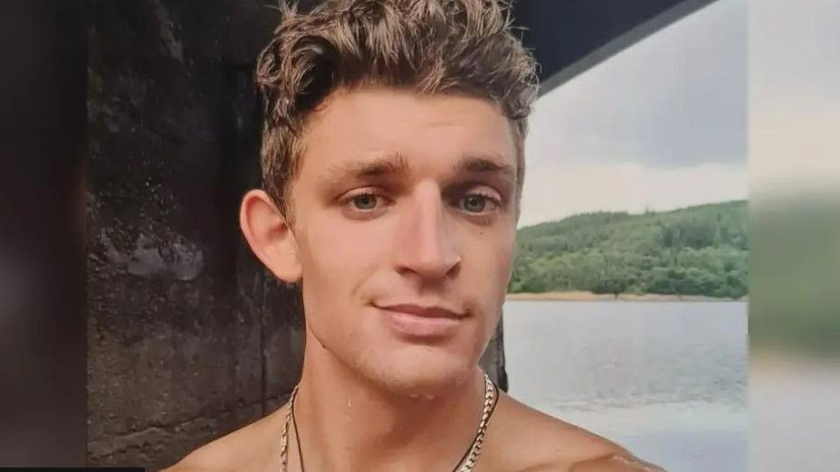 Haydn Griffiths smiles in this selfie, with the sea behind him and water seen on his face. He has blue eyes and short light brown hair and is wearing a gold chain.