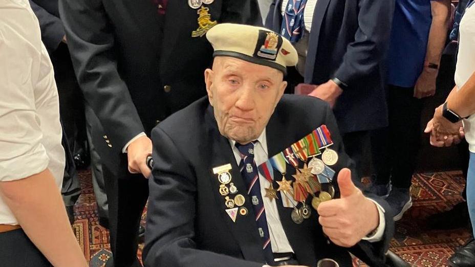 Dougie is wearing a cream coloured beret and a dark clue suit and tie, adorned with medals.  He is sticking his thumb up and sitting in his wheelchair.