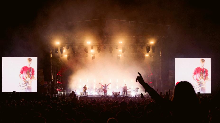 A shot from the crowd of Bloc Party on stage at Truck Festival. The lead singer's arms are aloft. The guitarist is pictured on the big screens.