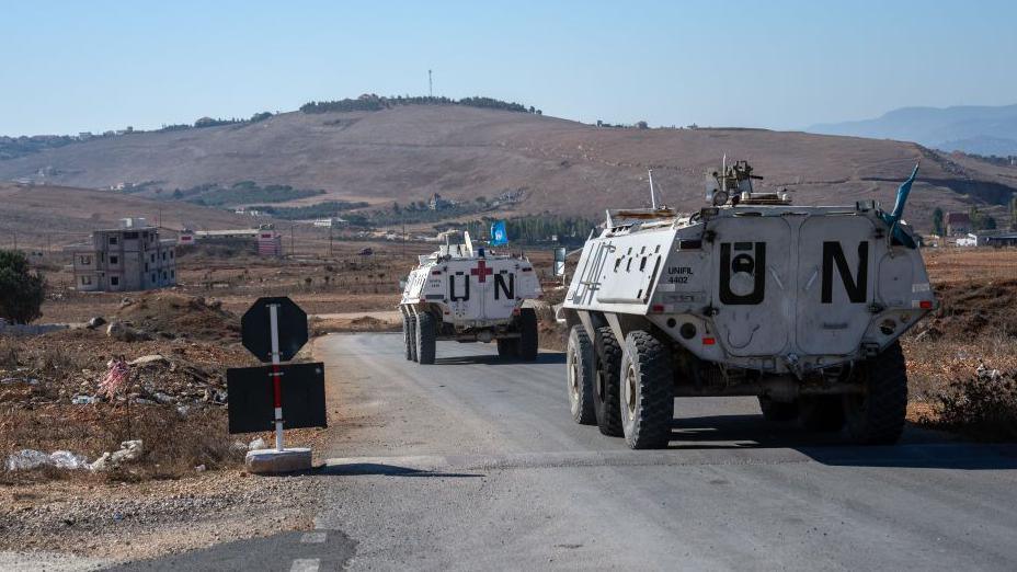 Two armoured white trucks, with UN painted on the back are driving towards a barren hill. A blue UN flag can be seen flying on the tops of the trucks.