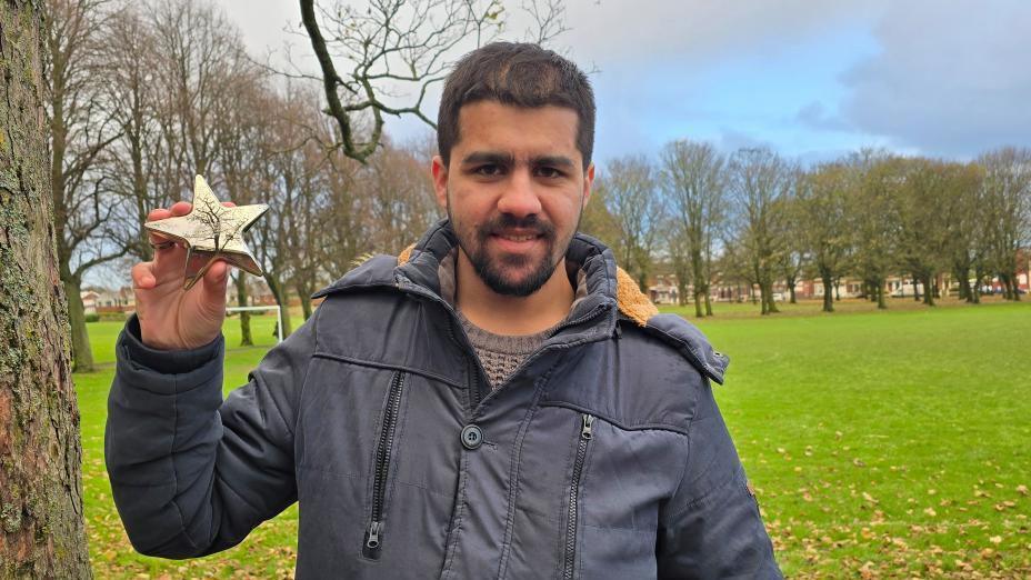 A young man stands in a green park wearing a navy jacket. He is holding a gold star trophy which is a Cancer Research UK Star Award for young people. 