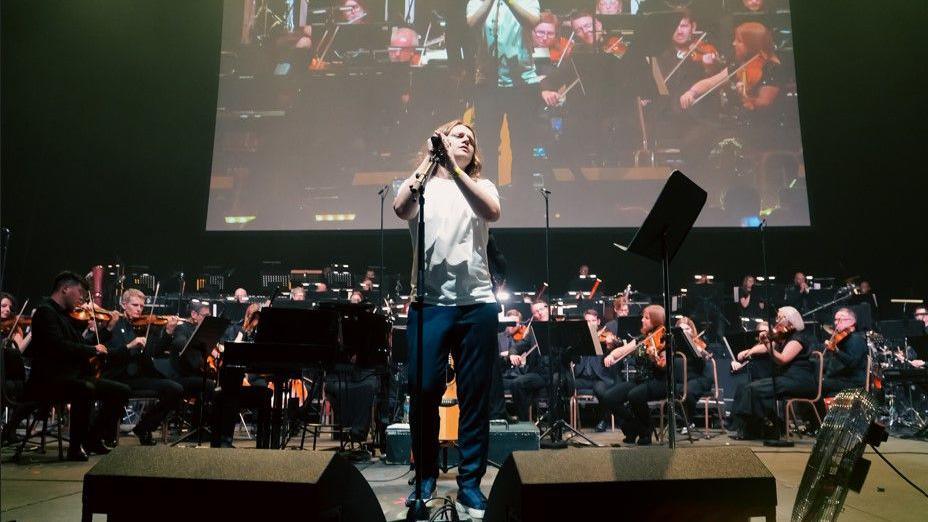 Jamie Webster is wearing jeans and a white T-shirt as he sings on stage, with the orchestra behind him. An image of him and the orchestra can be seen on a large screen behind them.