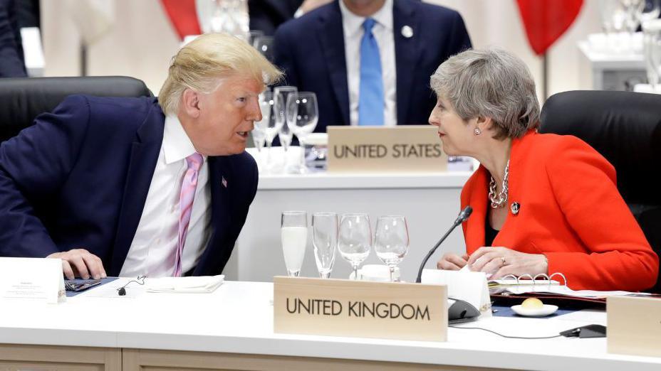 Donald Trump leans to speak to Theresa May, both seated at a G20 summit