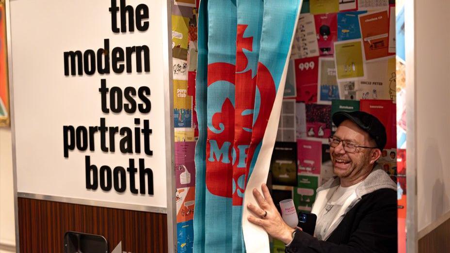 A man wearing a baseball cap and glasses is smiling as he looks through the blue curtain of the photo booth, which has 'the modern toss portrait booth' written on it. There are multiple Modern Toss cartoons pasted inside the booth.