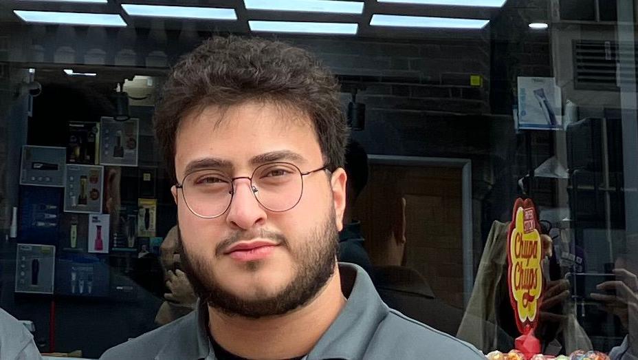 Ibrihim Gilgil, with beard, glasses and grey shirt, standing in front of his barber's shop