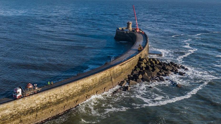 The new copper-coloured dome being transported on a reversing flat-bed lorry along the curving pier. A red crane has been set up by the lighthouse.