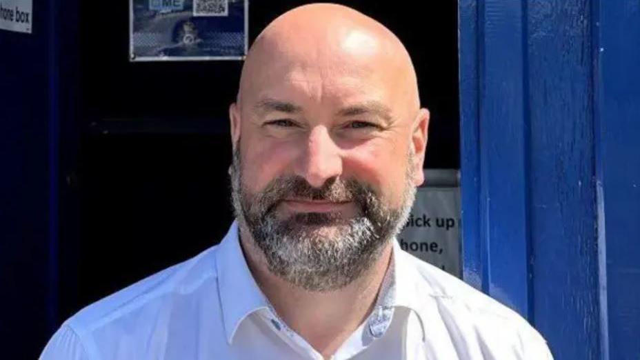Marc Jones wearing a white shirt and smiling as he stands in front of a blue-coloured small booth. He is bald and has a dark-coloured, but greying beard.
