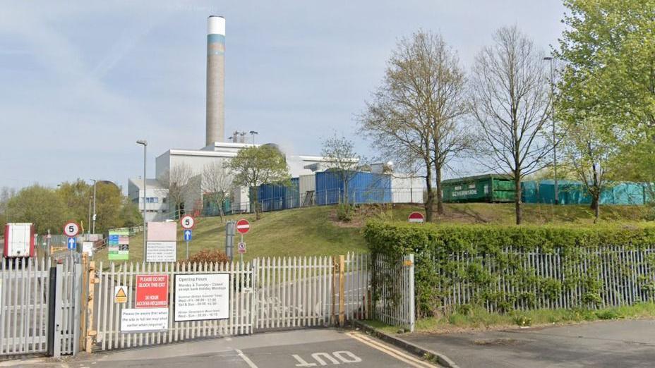 Closed metal gates and a fence surround the incinerator site, with grass beyond and the grey incinerator itself and the tall chimney above it