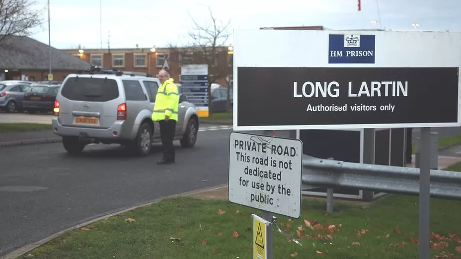 A Security guard wearing a high-vis jacket admits a silver vehicle into Long Lartin jail, Worcestershire