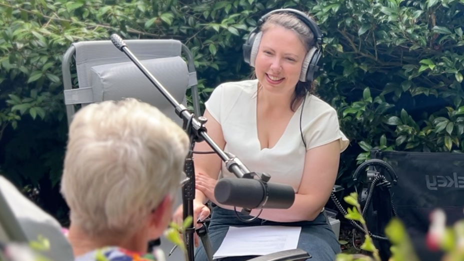 An elderly woman speaks into a microphone operated by a younger woman wearing headphones and smiling. They are both sitting on chairs in a garden.