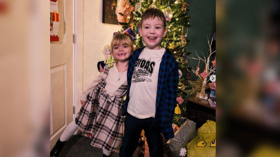 Jaxson hugging his sister, Bella. They are both smiling to camera with a Christmas tree in the background.