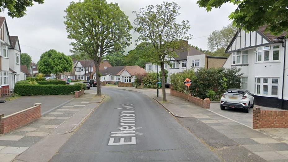 A leafy suburban street with detached houses on either side. There are green trees in bloom.