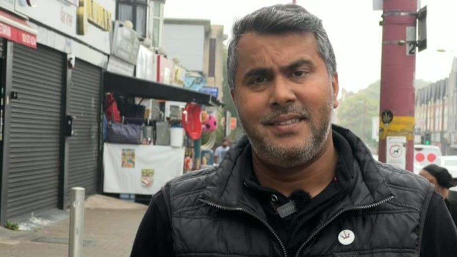 Bob Balu looking towards the camera with shops behind him. Mr Balu is wearing a black jumper and sleeveless jacket. One of the shops behind him has bags and other items for sale outside and another shop has closed shutters. 