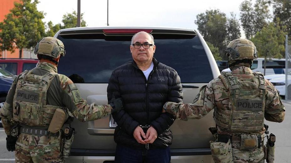 Osiel Cárdenas Guillén, wearing handcuffs, is held by two police officers in camouflage gear. Cárdenas is wearing a black puffer jacket and black-rimmed glasses. He is looking straight at the camera.