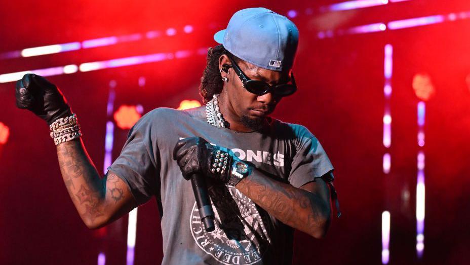 Offset performing in 2024. He wears a blue cap twisted back, dark sunglasses, a grey Ramones band t-shirt and studded black leather gloves. He looks down, holding his hands out to his right. The staging behind him is lit red. 