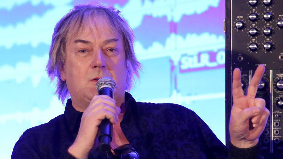 Geoff Downes, who has medium-length white hair, holding up two fingers in a V sign with his left hand and holding a microphone with his right