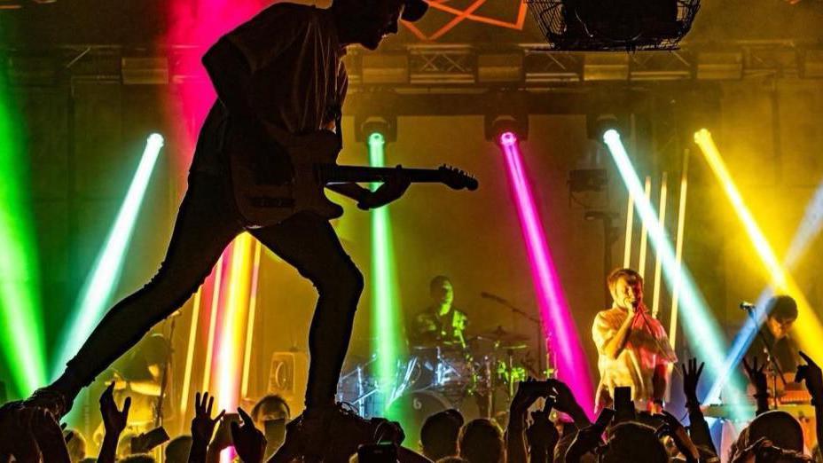 Band perorming on stage with a yellow and pink lighting background 