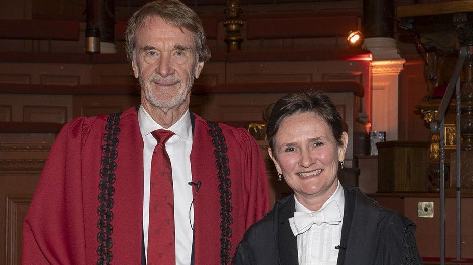 Sir Jim and Prof Tracey stood next to one another at the ceremony. Sir Jim is wearing a red tie and a red gown, Whilst Prof Tracey is wearing a white bow tie and a black gown.