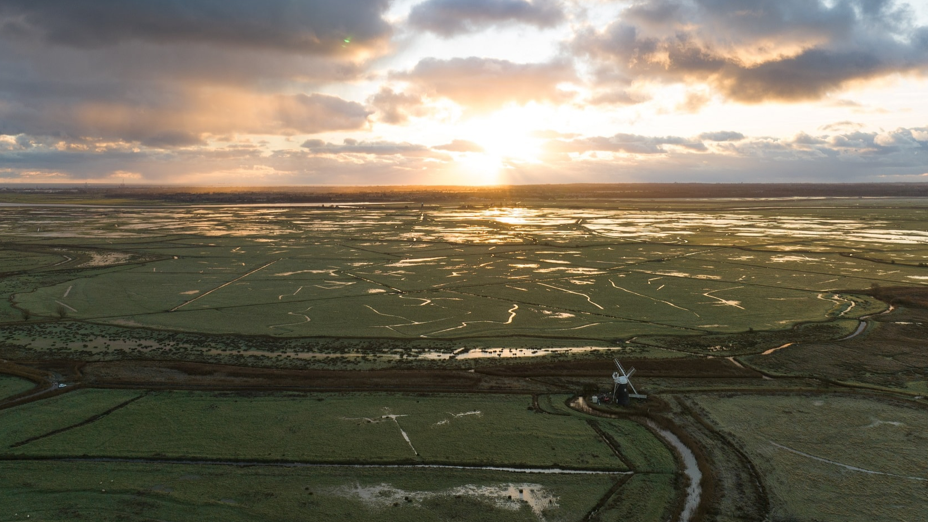 Aerial image of Halvergate marshes during a winter sunrise, showing Mutton's Mill (windmill) in the foreground and a waterlogged flat green landscape.