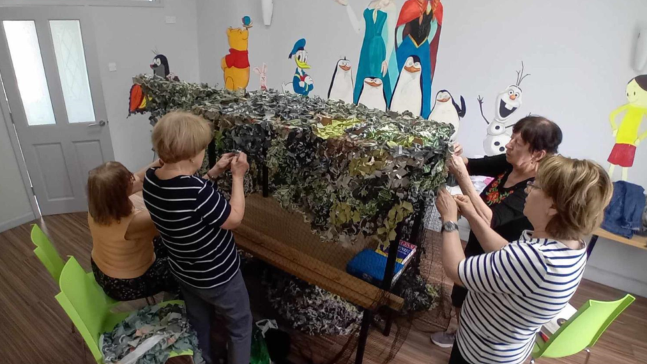 A group of four women standing around a table, working on attaching bits of brown and green fabric to what looks like a fishing net.