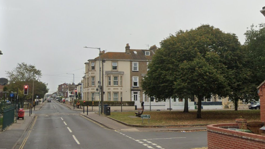 A town road is shown with a junction to another road to the right and a small green with trees on in the middle, with traffic lights on a crossroads to the left and houses in the background.