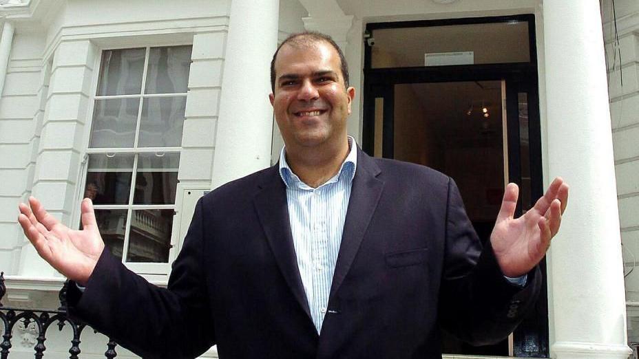 Sir Stelios Haji-Ioannou: A man with dark hair wearing an open-necked, blue-and-white-striped shirt and dark jacket. He has his arms outstretched and is standing outside the entrance to a property.