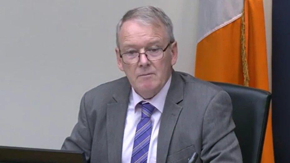 Brian Stanley is sitting in a committee. He's wearing black glasses and a grey suit with a white shirt and a purple tie. The Irish tricolour flag is behind him.