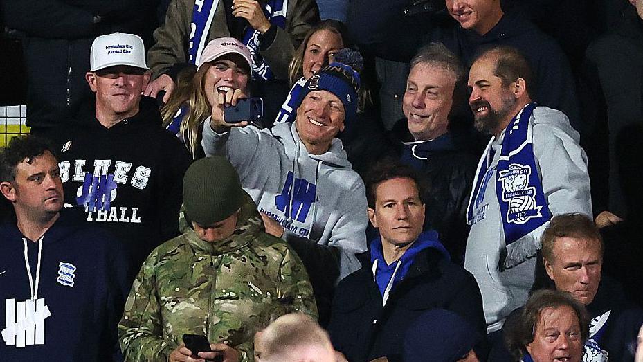 Tom Wagner in a grey Birmingham hoodie and bobble hat takes a selfie in the away end at Wrexham