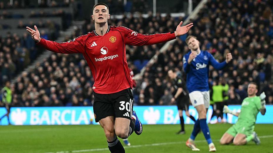 Benjamin Sesko runs away with his arms outstretched in celebration after scoring for Manchester United against Everton. Everton defender Jarrad Branthwaite and goalkeeper Jordan Pickford are out of focus in the background looking frustrated.