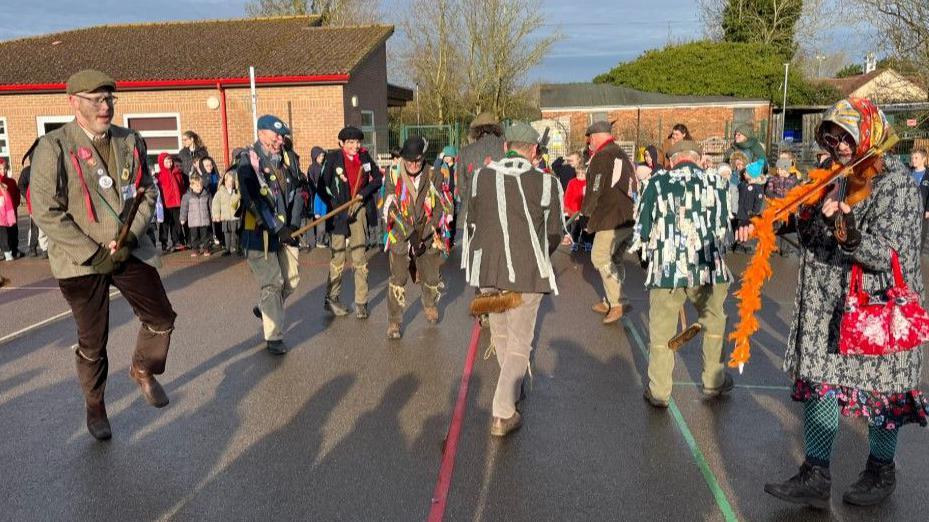 Molly dancers can be seen performing in front of a group of school children and parents or teachers. The dancers are dressed in old jackets and have string tied around their trousers. They are all wearing hats and some have brooms in their hands