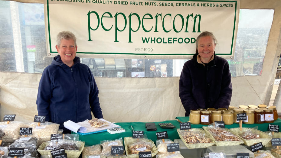 Two women at the back of a stall selling nuts and seeds smile at the camera