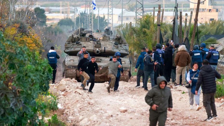 An Israeli tank rolls across a muddy and rocky road with several young Palestinians running away from it just in front and a huddle of journalists gathered on one side