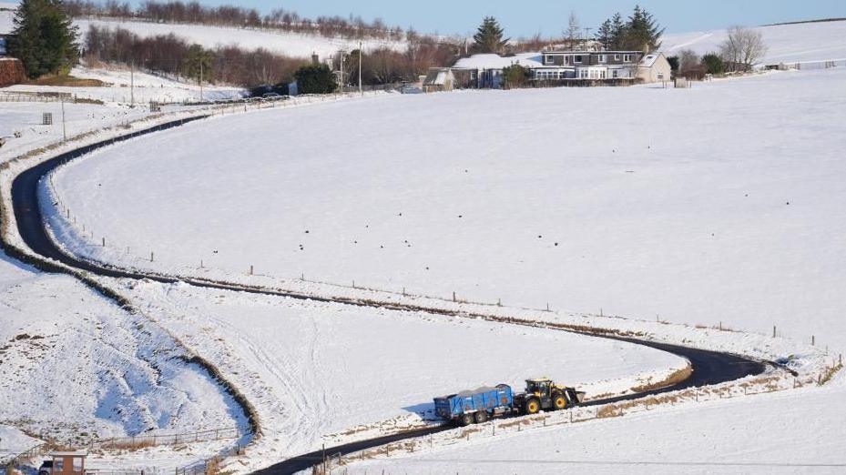 Snowy fields near Heriot, Scottish Borders, featuring a winding road on which a tractor makes its way with a loaded trailer