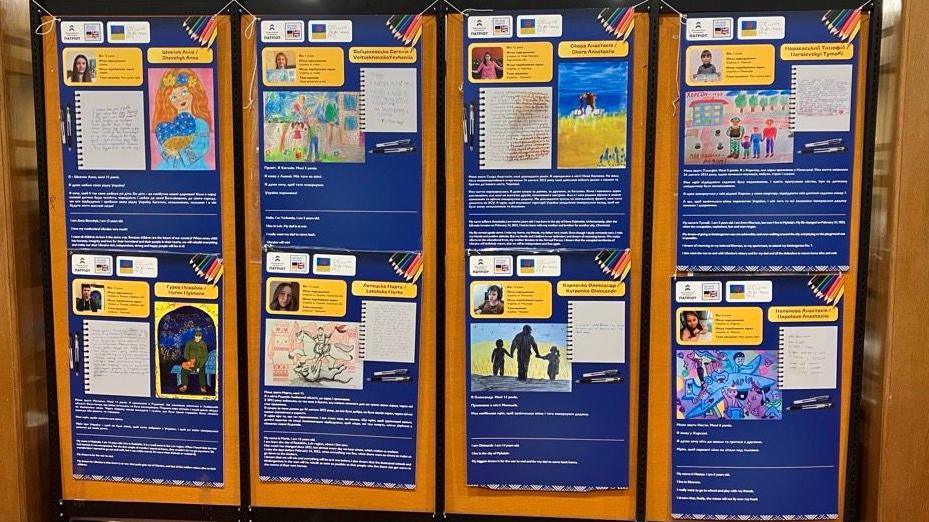 A display board featuring eight placards which showcase drawings and letters created by Ukrainian children. One photo shows the silhouette of a man walking into the sunset holding the hands of two children. 
