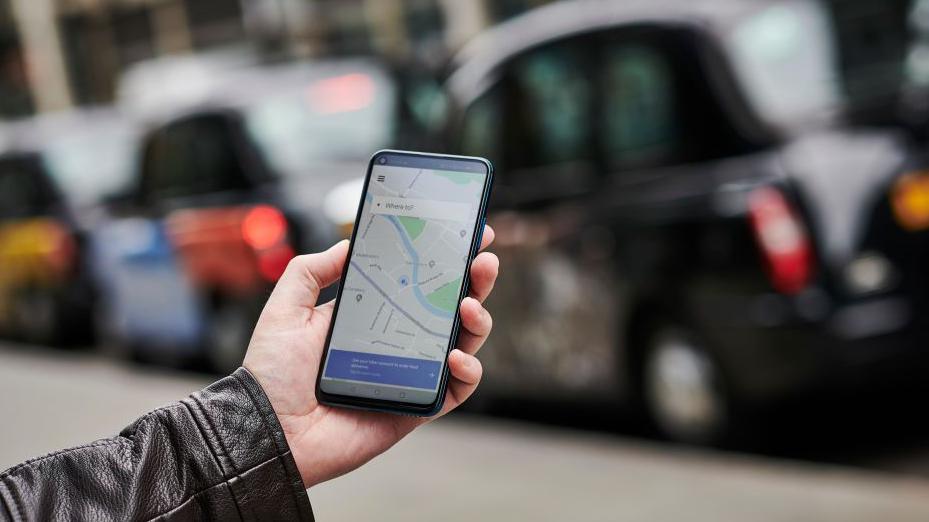 A hand holding a mobile phone which is on a map app. The arm shows the person is wearing a black leather jacket and behind their phone is a line of black taxis.