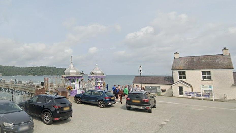 A google street view image shows a beige house on the  waterfront to the right of Garth pier, which has two small circular buildings either side of the start of the pier, which stretches out over the sea towards an greenary covered island in the distance. 