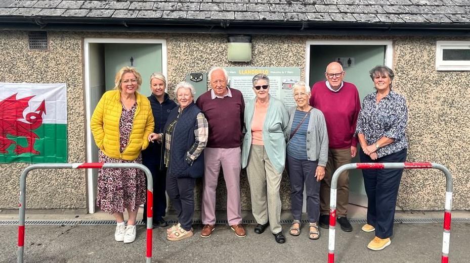 A group of eight volunteers stand outside a pebble dash toilet block. They are mostly elderly and a mix of men and women. On the toilet block wall is a large Welsh flag. The toilet block has two doors, one for men and one for women, and a grey slate roof, with two railings in front of it. The woman far left has a leopard print dress in the colours of pink and cream, and white trainers, over her dress is a yellow puffer coat, she has gold-framed glasses and short blonde hair. Next to her is a woman with blonde hair tied up and a blue coat and trousers. Next to her is a woman with a plaid shirt with a blue gilet over it, and blue jeans. She has cream Birkenstocks on and short grey hair. Next to her is na man with a dark pink jumper over a pink shirt, and pink trousers with brown shoes, he has white hair, and a receding hairline. Next to him is a woman with shades on and short grey hair, she is wearing a peach top with a light-green cardigan, and cream trousers. Next to her is a woman with a blue striped t-shirt and grey cardigan, she also has blue trousers on and the brown strap of a handbag across her shoulder. Next to her is a man with a pink jumper and brown trousers, he is bald. And on the right is a woman with her arms in front of her, she has a flowery dark blue top on with dark blue jeans. She has short grey hair.