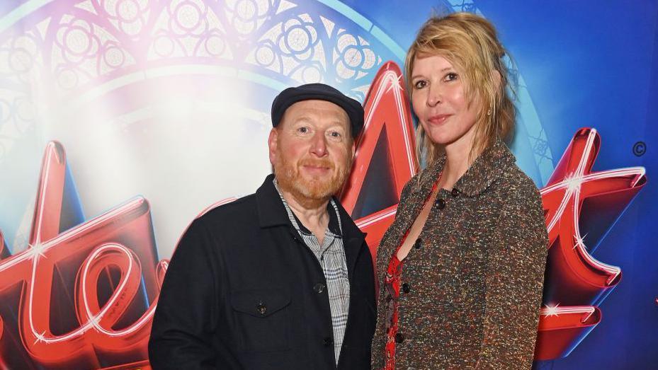 A photo of a man and woman in front of a Sister Act logo. The man is wearing a black flat cap, and a black jacket. He has a ginger beard. The woman on the right has blonde hair and wears a colourful jacket. They both look at the camera.