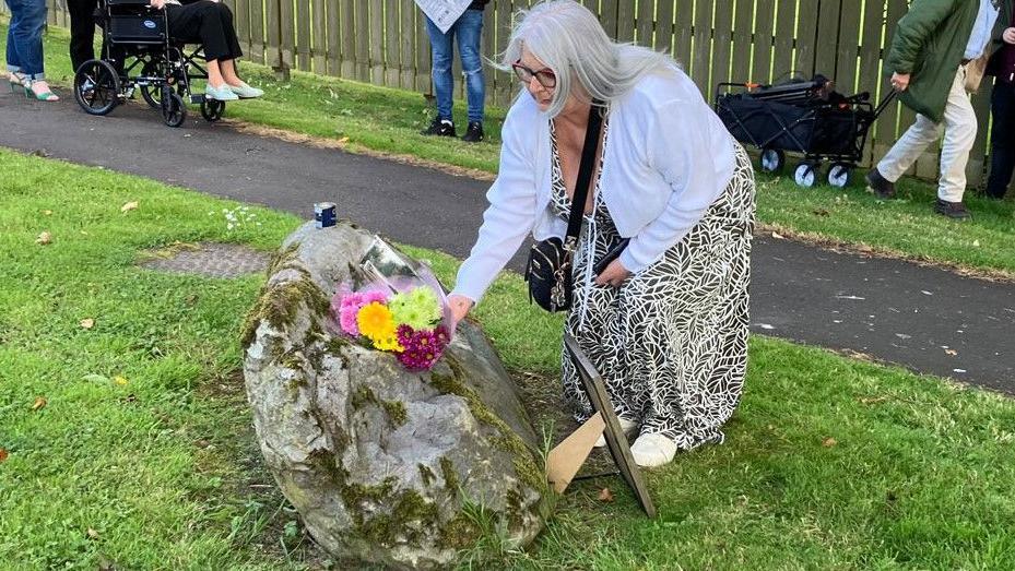 Kathleen Arkinson, wearing a black and white printed dress with a white cardigan, lays flowers by the memorial stone in Castlefinn Park